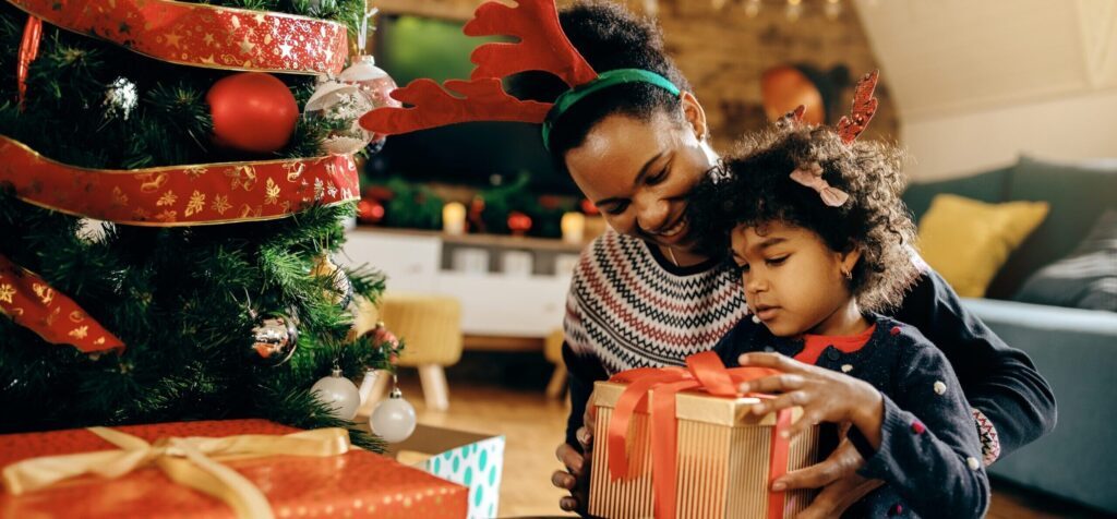 Little girl and her mother open Christmas presents by the tree.